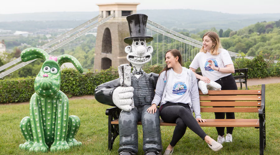 Two women sitting on a bench with Wallace & Gromit statues in front of the Bristol Suspension Bridge.