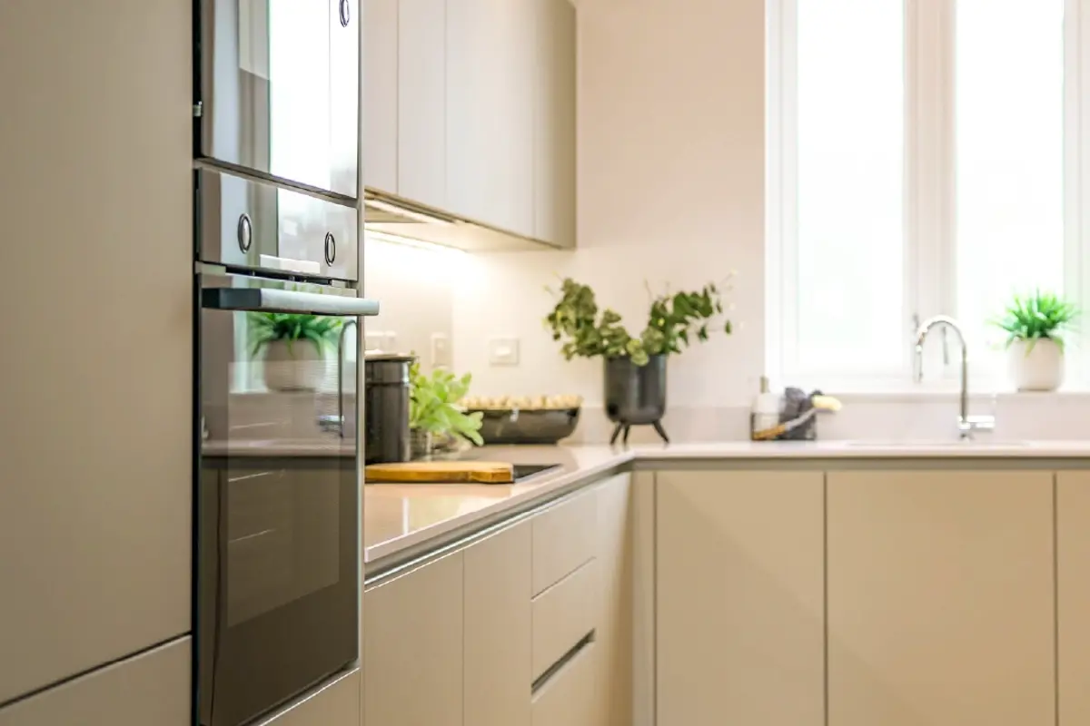 Modern kitchen with sleek cabinets, built-in oven, fresh plants, and natural light from large windows. Minimalist and inviti