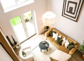 A stylish, airy living room featuring a tan sectional sofa, cozy chairs, a round black coffee table, and large patio doors to a garden.