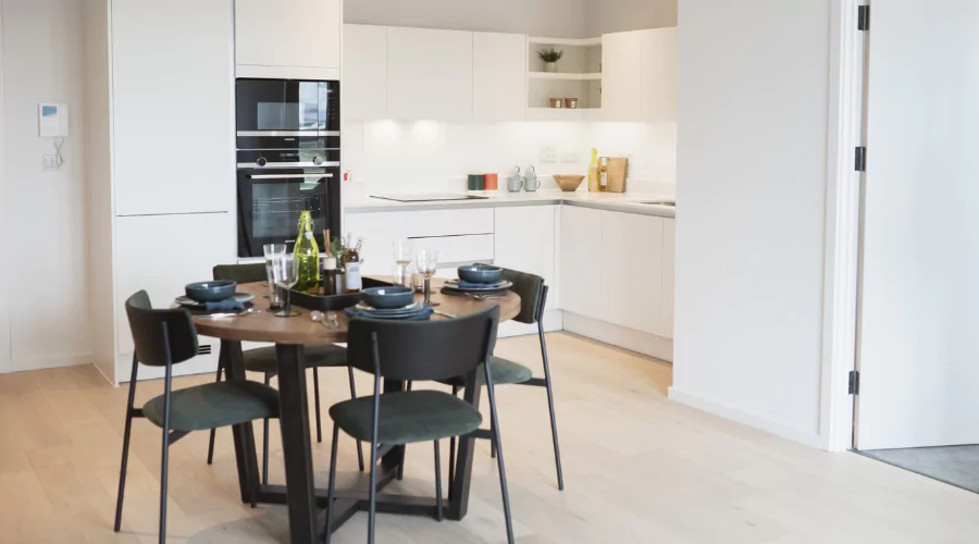 Pre set dining room table with four black chairs placed in front of lit up modern and clean kitchen.