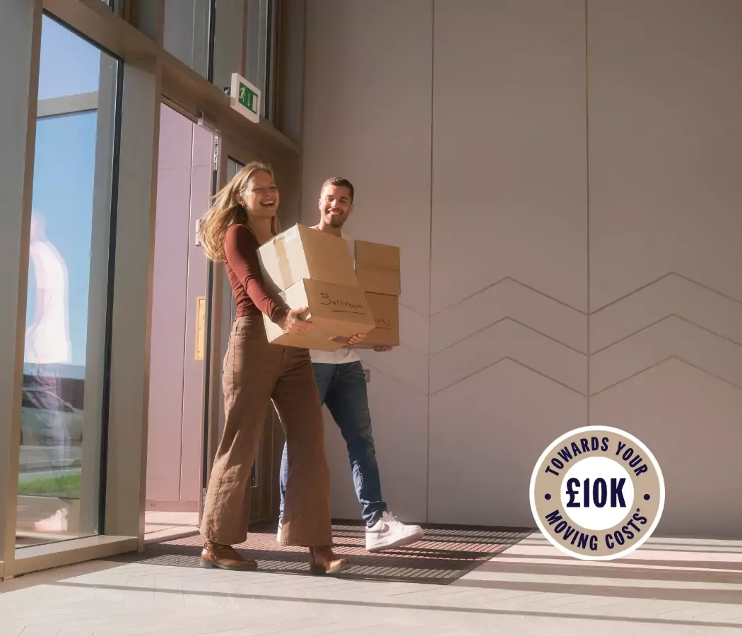 A smiling couple carrying moving boxes enters the foyer of the Dials apartments at Brabazon. A "£10K towards your moving costs." sticker overlays the image.