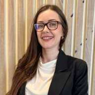 Close up photograph of a smiling young woman with long brunette hair, glasses and a black blazer