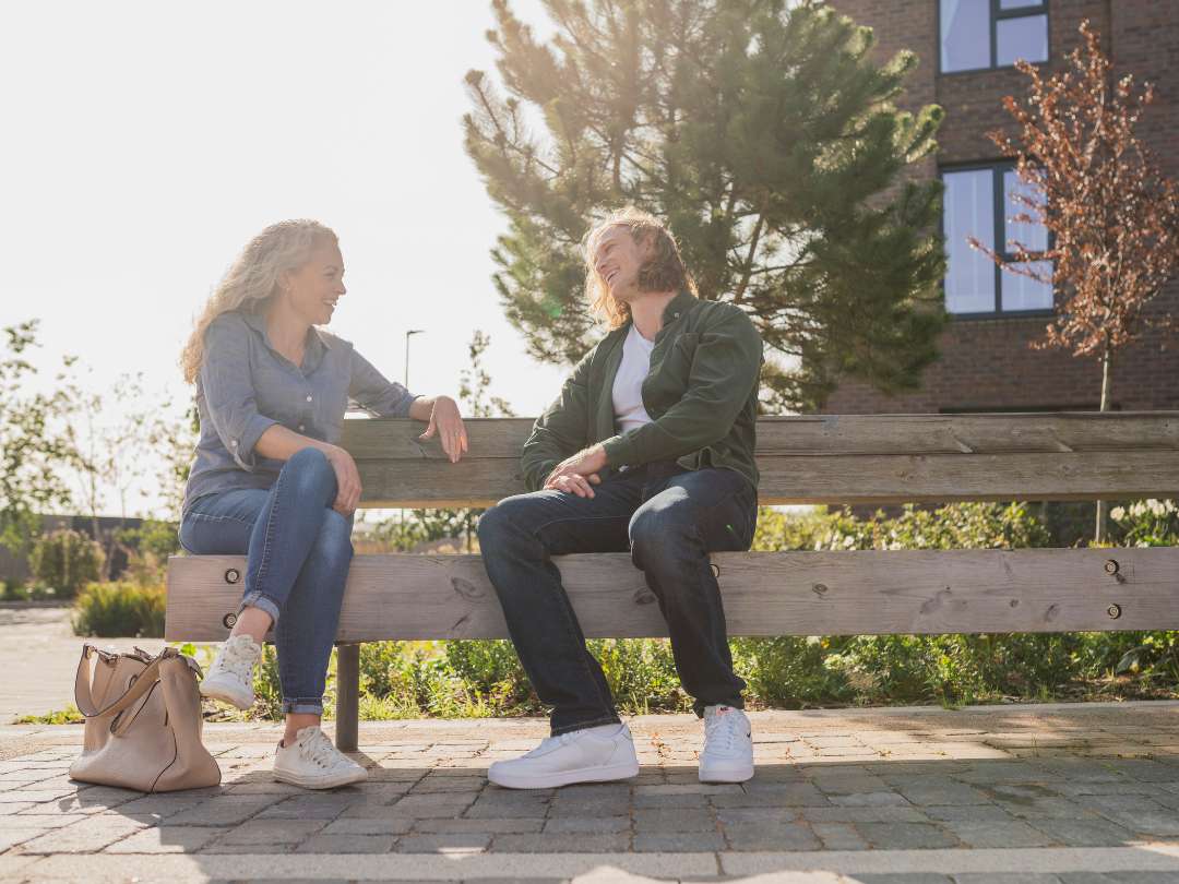 Older female and young male laughing and smiling on a wooden bench in the sun at The Hangar District. 